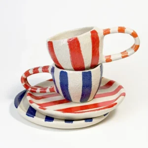 Two striped ceramic mugs with matching round plates, one set in blue and red stripes, the other in red and orange stripes.